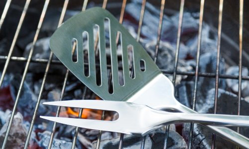 Close-up of a spatula and grilling fork resting on the edge of a lit charcoal grill, with glowing coals visible beneath the grate.